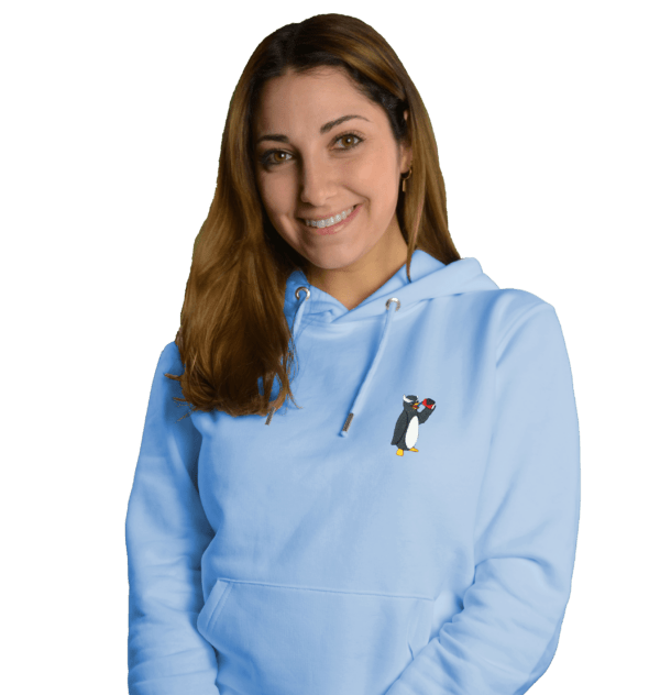 Eine lächelnde Frau trägt einen hellblauen nachhaltigen Hoodie mit einem kleinen Pinguin-Logo auf der linken Brust.